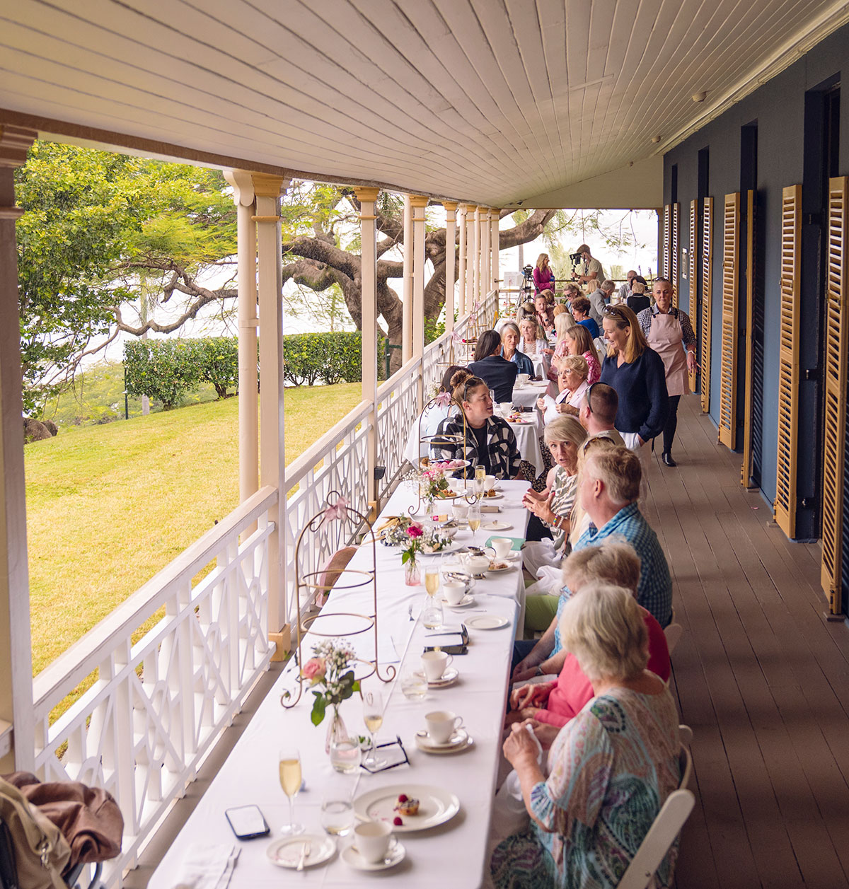 Covered verandah with white clothed tables along it and people seated at the tables looking out rowards a park and river view