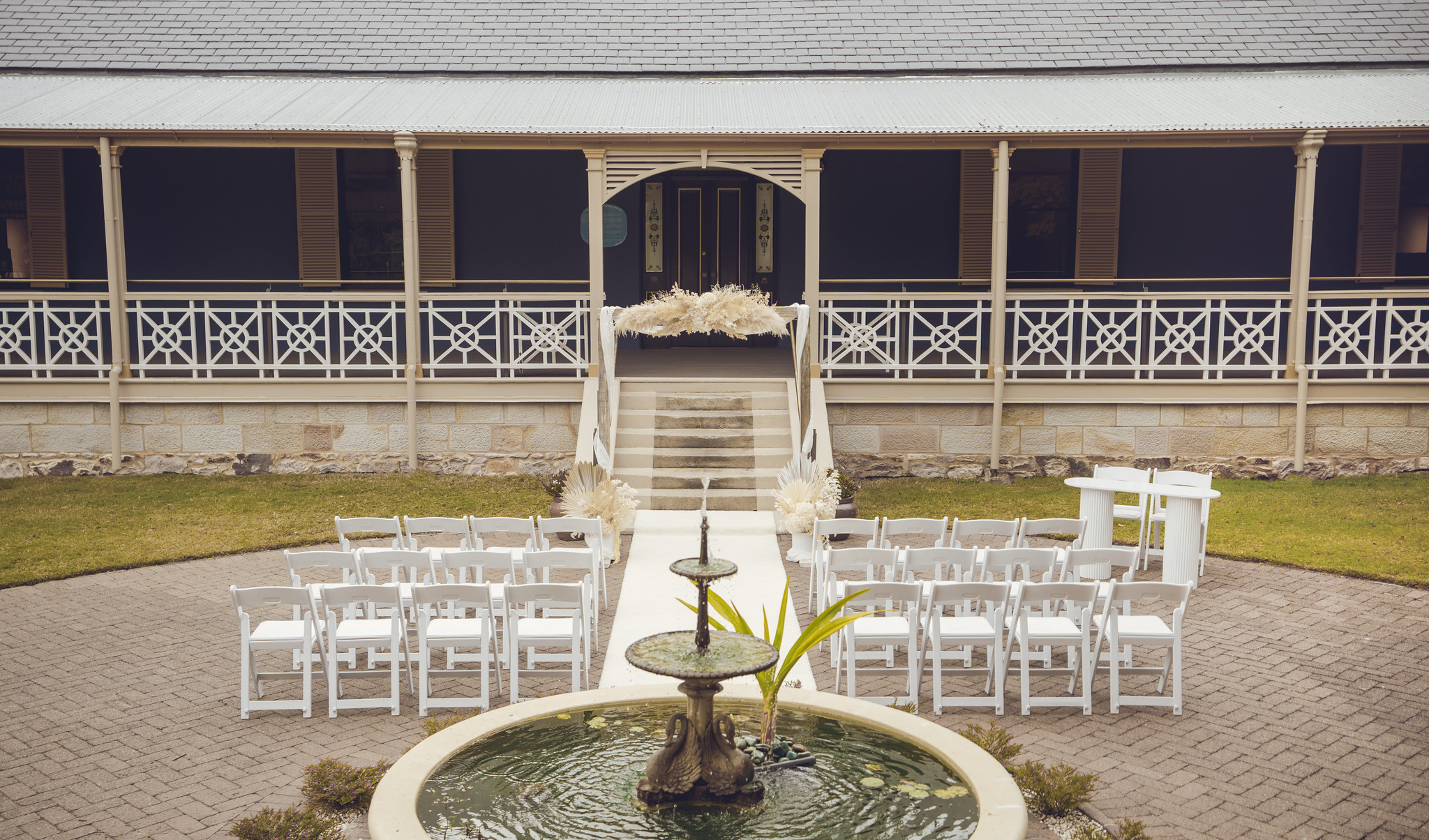 Circular stone fountain filled with water at the base whote wedding chairs in front and wedding arch with white dried flowers house verandah behind