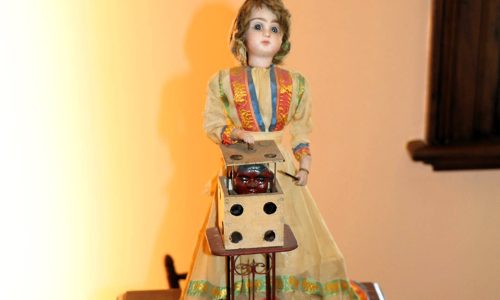 Porcelain Doll wearing a dress and bonnet with a dice shaped box in front showing a devil head popping out of the box
